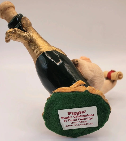 A collectible figurine of a pig supporting a champagne bottle, with the pig's snout and back visible, on a plain light background. The base features a green velvet-like finish with a white label that reads 'Piggin' Piggin' Celebrations by David Corbridge Hand Made 1999 DDCWALCWSL'.