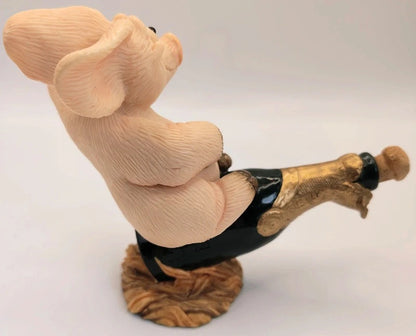 A pig figurine wearing a black top and holding a champagne bottle, mounted on a textured base, photographed against a plain light gray background.