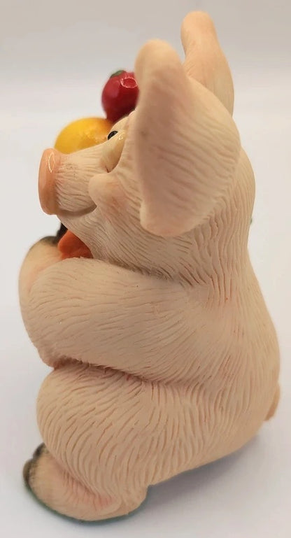 A small sculpted pig figurine shown from the back, holding a cluster of colorful fruits, photographed against a plain light background.