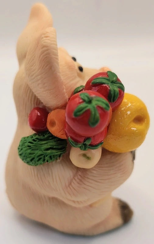 A small sculpted pig figurine holding a bunch of colorful vegetables, including red tomatoes, a yellow pear, green broccoli, and an orange carrot, on a plain light background.