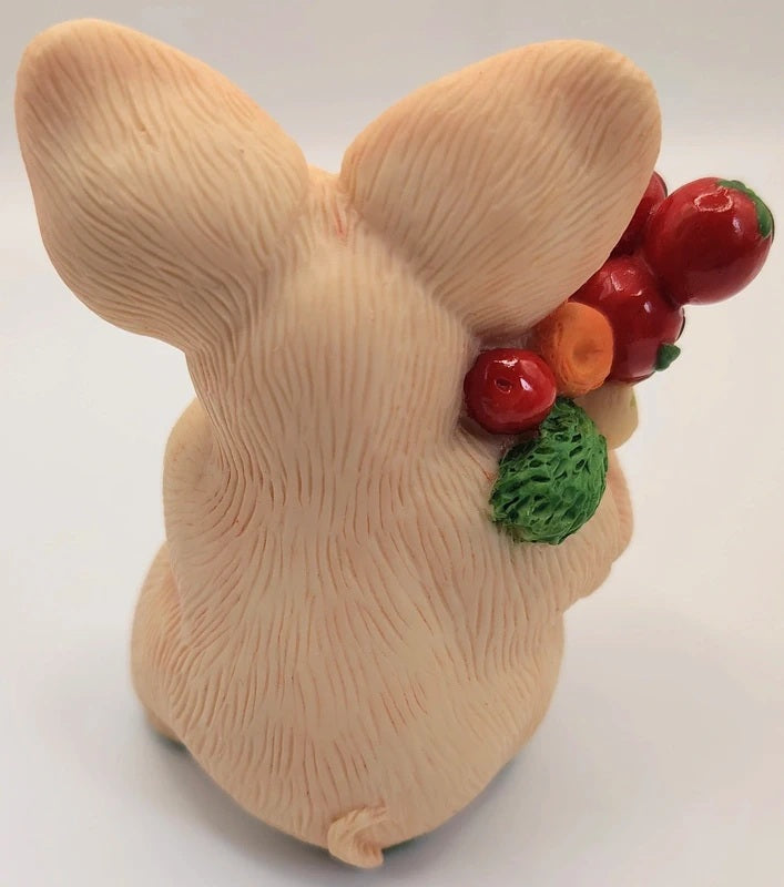 A beige bunny figurine shown from the back, holding a small bunch of red berries and green leaves, photographed on a plain light background.