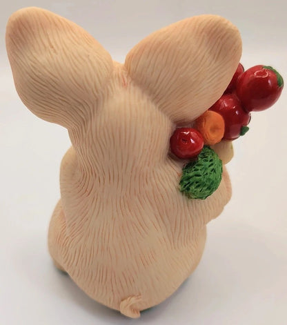 A beige bunny figurine shown from the back, holding a small bunch of red berries and green leaves, photographed on a plain light background.