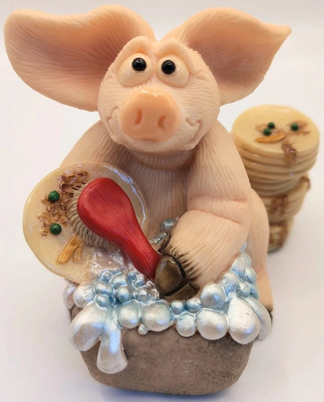 Piggin Dishes Figurine hand made by David Corbridge in 1999. A whimsical pig figurine sitting in a tub of soapy water, holding a red brush. The pig has large pinkish ears and a pink snout, with a small stack of round decorative items behind it on a plain white background.