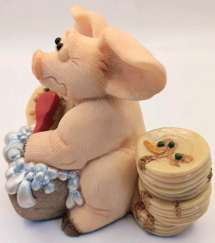 A pig figurine sitting on a stone-like base, holding a red heart-shaped object, with a stack of round plates beside it. The figurine appears to be made of a glossy resin or similar material, shown against a plain white background.