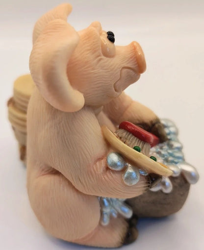 A small sculpted figurine of a pig holding a basket or tray with shiny silver beads and a red accent, photographed against a plain light background.