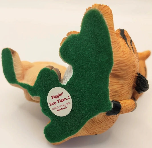 A handmade pig-shaped tiger figurine shown on its back, featuring orange-brown textured fur, a green felt back, and black paw pads. A white circular sticker on the back reads 'Piggin' Easy Tiger.