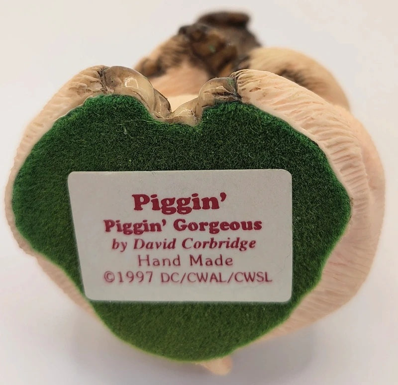 A close-up of the back of a Piggin' figurine by David Corbridge, featuring a beige textured body, a green felt patch, and a white label with red text.