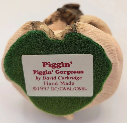 A close-up of the back of a Piggin' figurine by David Corbridge, featuring a beige textured body, a green felt patch, and a white label with red text.