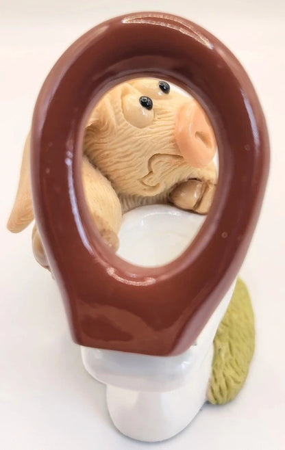A ceramic figurine of a pig peeking out from behind a brown toilet lid, mounted on a white ceramic base.