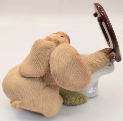 A ceramic figurine of a beige pig sitting on a white toilet with its brown front paw resting on the open lid, set against a plain white background.