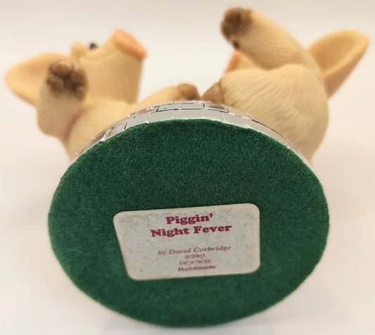 A close-up of the circular base of a handcrafted ornament featuring a beige pig figurine lying on its back, with a green felt top and a silver-wrapped edge. The base has a white label reading 'Piggin' Night Fever' by David Cartridge.