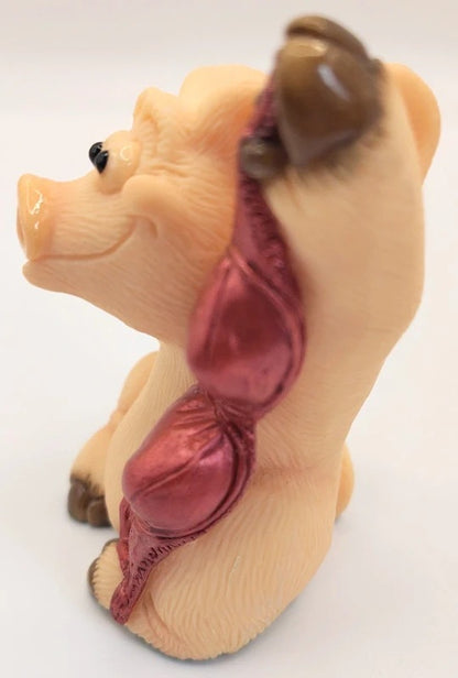 A novelty pig figurine wearing a metallic pink and purple bra, set against a plain white background.