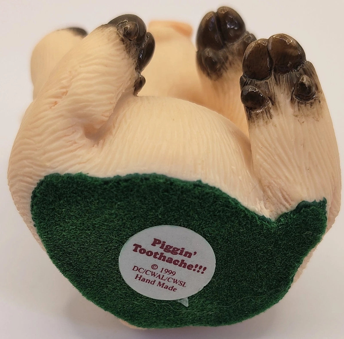 A vintage resin pig figurine shown from underneath, featuring cream-colored legs with dark brown hooves, a green textured belly, and a white sticker that reads 'Piggin' Toothache!!!'.
