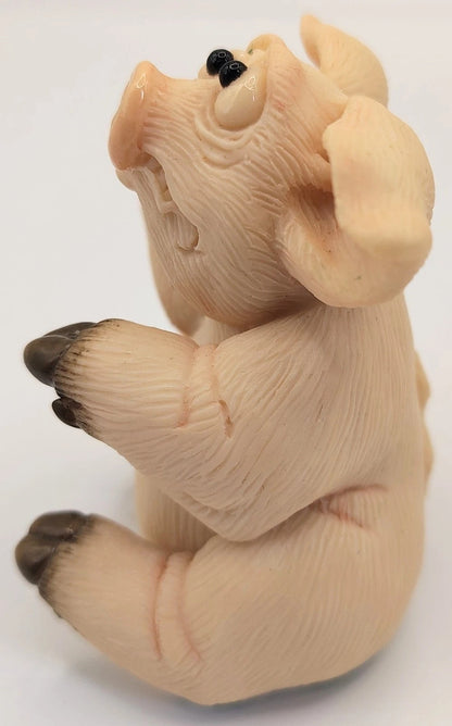 A small sculpted pig figurine in a seated pose, shown against a plain white background. The pig has a pale peach-beige body with darker brown hooves and black eyes.