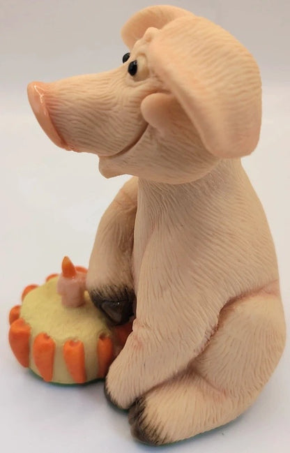 A beige figurine of a pig sitting next to a small orange and yellow cake, photographed against a plain white background.