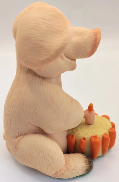 A beige pig figurine sitting beside a small yellow cake with orange frosting and candles, photographed against a plain light background.