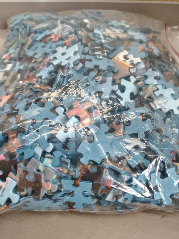A clear plastic bag filled with many jigsaw puzzle pieces, photographed from above on a light tan surface.