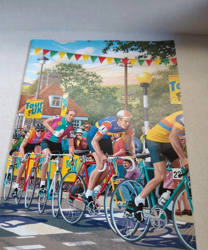 A large rectangular wall art print or poster depicting a colorful cycling race scene, mounted against a light beige wall. The artwork shows multiple cyclists in bright team jerseys riding road bikes, with houses, trees, and festive bunting in the background.