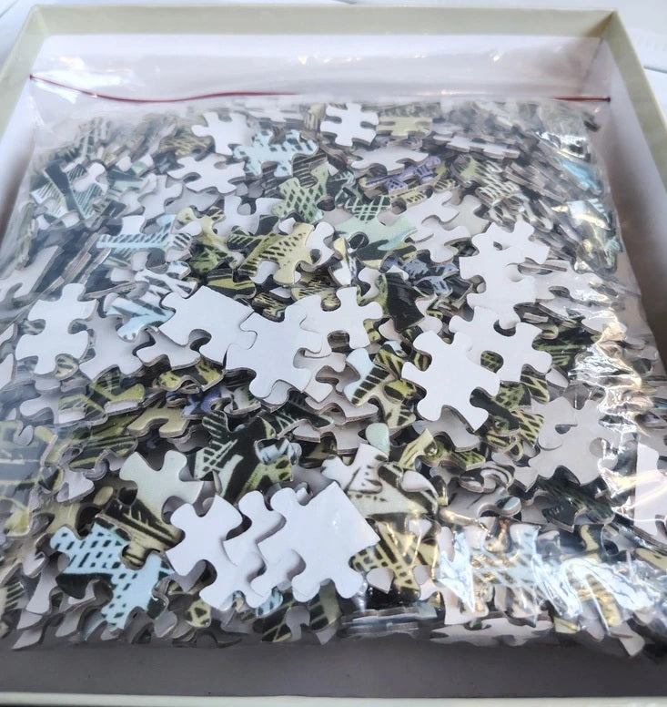 A box of jigsaw puzzle pieces inside a clear plastic bag, showing a multicolor abstract pattern with white puzzle pieces mixed throughout.