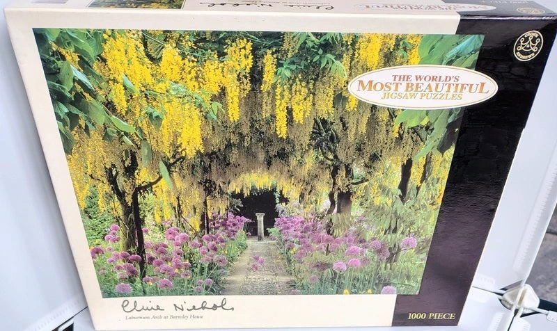 The Worlds Most Beautiful Jigsaw Puzzles Clive Nichol Laburnum Arch at Barnsley House 1000 Piece Jigsaw Puzzle. A 1000-piece jigsaw puzzle box featuring a garden scene with yellow wisteria hanging over a path and pink flowers in the foreground. The box has a white border and a black side panel with branding and product text.