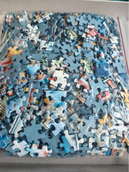 A large pile of colorful jigsaw puzzle pieces inside a clear plastic bag, photographed from above on a light gray surface.