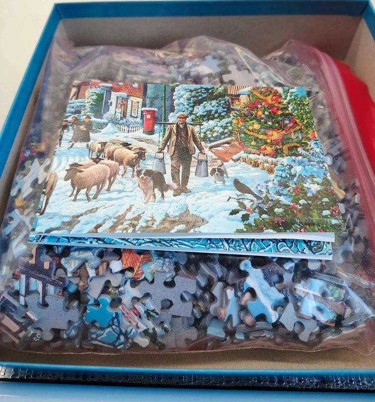 A completed winter-themed jigsaw puzzle in a blue box, with several loose puzzle pieces visible underneath a clear plastic bag. The puzzle depicts a snowy village street scene with people, sheep, dogs, and a decorated Christmas tree.
