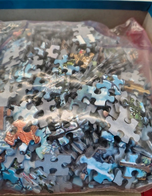 A bag of jigsaw puzzle pieces with a light blue and gray base and small multicolored printed details, photographed inside a box.