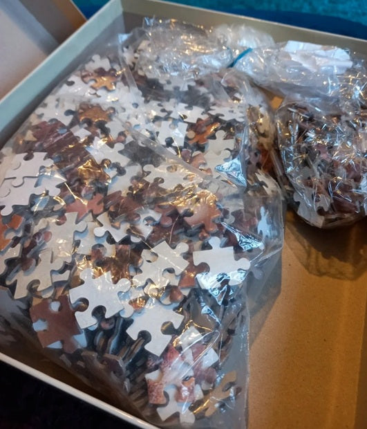 Jigsaw puzzle pieces in a box with some pieces outside, still in plastic wrap.