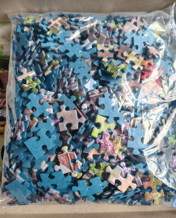 A clear plastic bag filled with assorted jigsaw puzzle pieces, many of which are blue, photographed from above on a light beige surface.