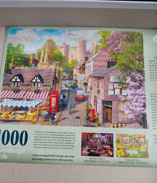 A printed 1000-piece jigsaw puzzle package or puzzle insert featuring a colorful village street scene with stone towers, red buses, market stalls, and blooming pink trees. The package includes product text and small thumbnail puzzle result photos along the bottom.