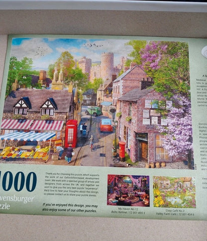 A printed 1000-piece jigsaw puzzle package or puzzle insert featuring a colorful village street scene with stone towers, red buses, market stalls, and blooming pink trees. The package includes product text and small thumbnail puzzle result photos along the bottom.