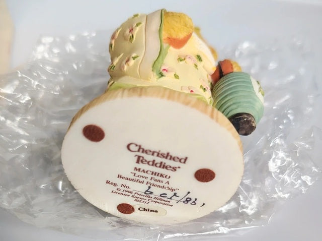 A vintage Cherished Teddies figurine of Machiko on bubble wrap packaging. The figurine features a cream-colored body with floral patterns, a green and white striped shirt, and a yellow-orange collar.
