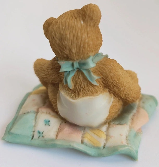 Teddy bear figurine with a bow sitting on a decorative pillow against a white background