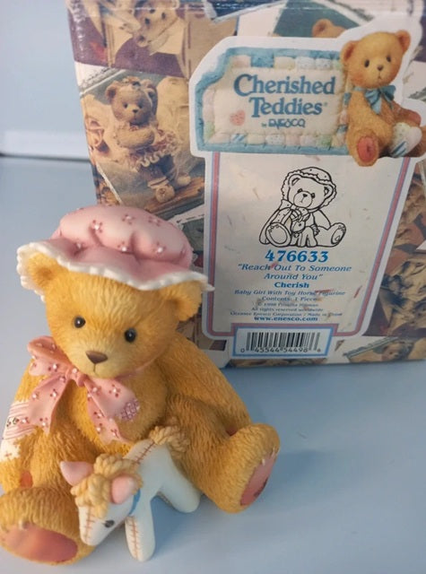 Teddy bear with a pink hat and scarf in front of a Cherished Teddies packaging.