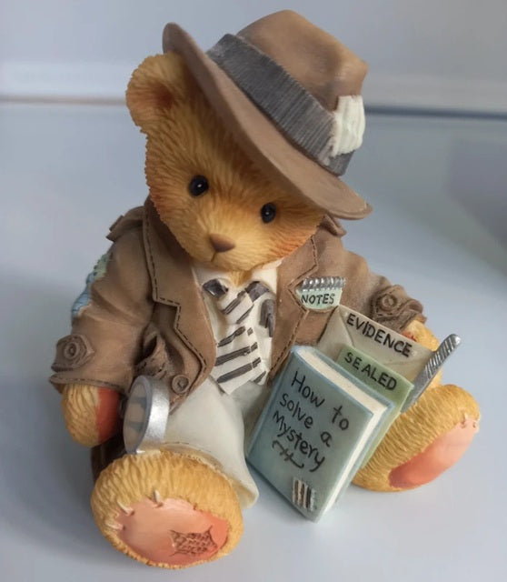 Teddy bear dressed as a detective with a book titled 'How to Solve a Mystery' on a white background