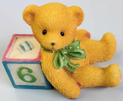 Yellow teddy bear with a green bow sitting next to a wooden block with numbers on a white background