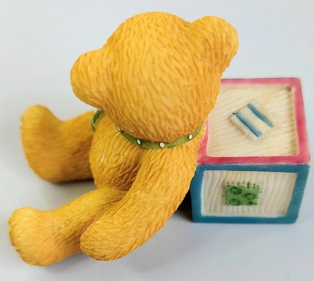 Yellow teddy bear with a green collar next to a wooden block set on a white background