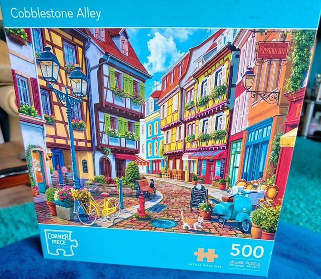 Box for a 500‑piece jigsaw puzzle titled “Cobblestone Alley,” showing a colourful European‑style street with bright buildings, flower boxes, a yellow bicycle, a blue scooter, two white cats and cobblestone paving.