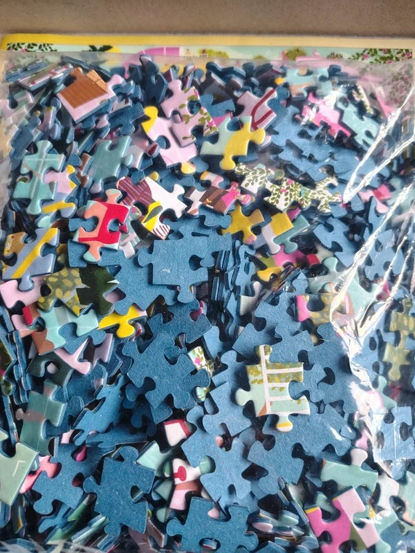 A clear plastic bag filled with assorted jigsaw puzzle pieces in many colors, including blue, pink, yellow, white, and green, photographed from above.