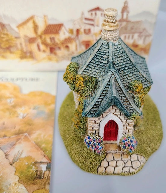 Small fairy house with a red door on a green base, with a scenic village background.