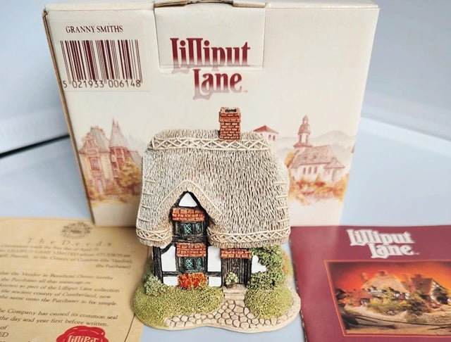 A Lilliput Lane collectible cottage figurine displayed in front of its original cream-colored packaging box, with a vintage insert card and a red booklet on a light blue surface.