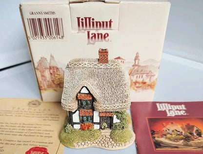 A Lilliput Lane collectible cottage figurine displayed in front of its original cream-colored packaging box, with a vintage insert card and a red booklet on a light blue surface.