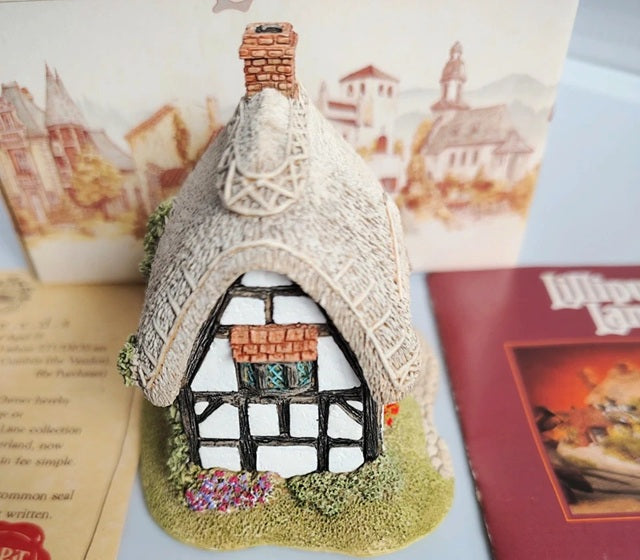 A small decorative cottage figurine with a thatched roof, black-and-white timber framing, and a brick chimney, displayed with printed cards or booklets in the background.