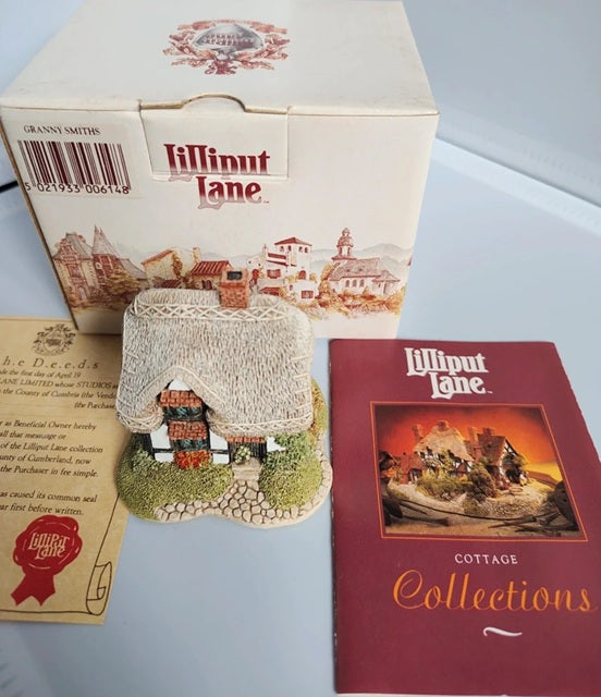 A Lilliput Lane Cottage Collection miniature village cottage figurine displayed alongside its original cream-colored packaging box, a printed booklet, and a red 'Cottage Collections' booklet on a light gray surface.
