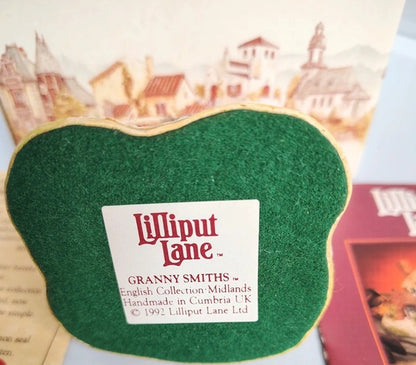 A green felt Lilliput Lane Granny Smiths cookie shaped plaque or ornament with a white paper label reading 'Lilliput Lane Granny Smiths English Collection Midlands Handmade in Cumbria UK 1992 Lilliput Ltd', shown against a printed book or card background.