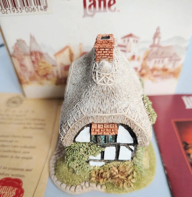A small decorative Lilliput Lane Granny Smiths thatched cottage figurine with a brick chimney, white-and-black timber framing, and a textured grassy base, shown in front of its printed packaging box.