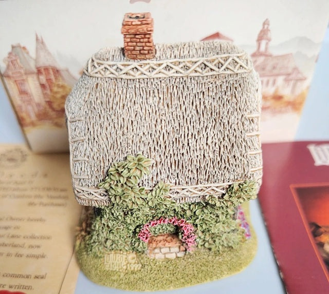A miniature decorative cottage figurine with a textured beige thatched roof, a small brick chimney, green mossy base, and a tiny pink flower archway, displayed among printed cards and books.