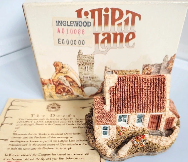 A miniature decorative model of Inglewood Cottage 'Lilliput Lane' featuring a detailed brick house with a stone wall and small garden, displayed in front of its illustrated cardboard box.