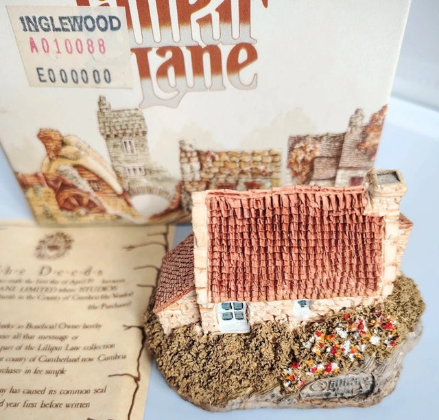 A miniature decorative model of a cottage in Sturton, Cumbria, UK, displayed in front of its printed product box. The model features a textured reddish-brown tiled roof, a small white window, and a rustic stone base labeled 'Sturton'.
