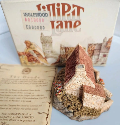 A miniature model of a cottage in Lilliput Lane called Inglewood, shown with its illustrated packaging box and an informational card on a light blue background.